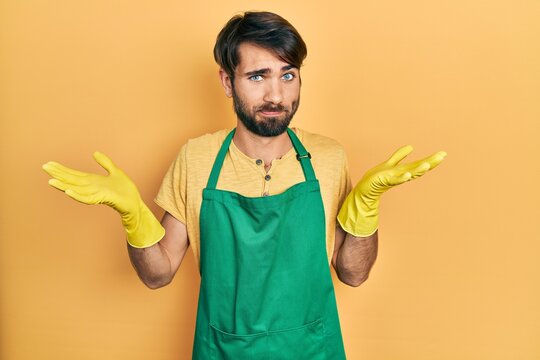 Young hispanic man wearing cleaner apron and gloves clueless and confused with open arms, no idea and doubtful face.
