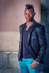Dressing in a black blazer, green pants, a young black man with mohawk hair is standing against the wall, confidently looking at you..