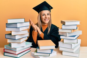 Young caucasian woman wearing graduation ceremony robe sitting on the table clapping and applauding happy and joyful, smiling proud hands together