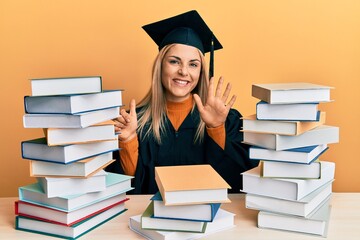 Young caucasian woman wearing graduation ceremony robe sitting on the table showing and pointing up with fingers number six while smiling confident and happy.