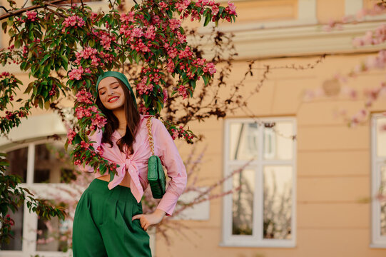 Elegant happy smiling woman wearing stylish green bandana, pants, pink shirt with knot, posing near blooming  tree. Spring, fashion, lifestyle conception. Outdoor portrait. Copy, empty space for text
