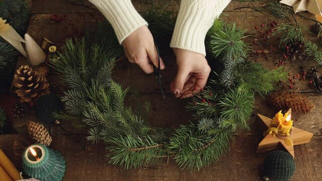 Making Christmas wreath. Top view of woman hands making Christmas wreath with fir tree and red berries on rustic wooden table with candles and pine cones. Holiday advent. Merry Christmas