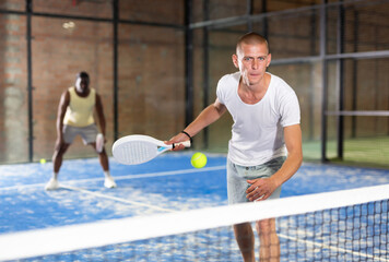 Portrait of concentrated paddle tennis player preparing to hit forehand to return ball on indoor court..