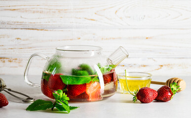Transparent glass kettle with strawberry and mint tea on white wooden background. Healthy organic fruit drink.
