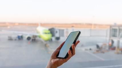 Close-up shot of female hand holding smart phone by window at airport. Area with airplane in background