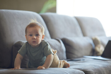 Candid portrait of cute baby boy crawling on sofa and looking at camera in cozy home interior, copy space
