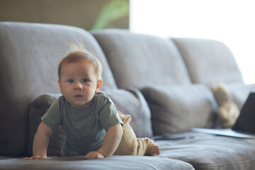 Fototapeta premium Candid portrait of cute baby boy crawling on sofa and looking at camera in cozy home interior, copy space