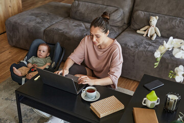 High angle portrait of modern mother working from home with cute baby boy lying in carrier, copy space