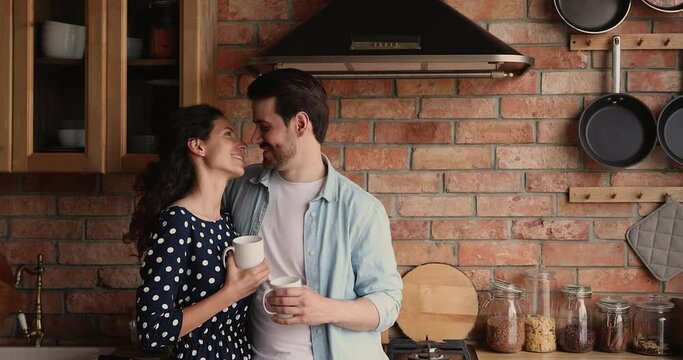 Loving young husband cuddling smiling beautiful wife in kitchen, enjoying pleasant conversation while drinking morning coffee together, spending leisure weekend lazy time communicating at own home.