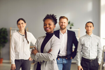 Happy aspiring black business woman in suit smiling at camera standing arms folded with team of workers behind. Portrait of ambitious female leader, executive, bank staff manager. Blurred background