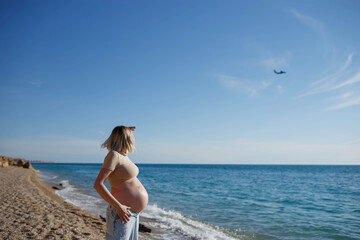 young pregnant girl by the sea looks at a flying plane