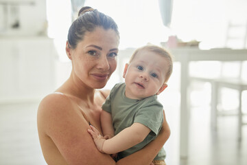 Candid portrait of freckled adult mother holding baby boy and looking at camera in cozy home...