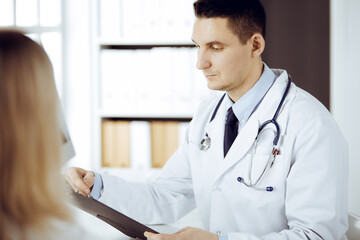 Friendly male doctor and patient woman discussing current health examination while sitting in sunny clinic. Perfect medical service in hospital