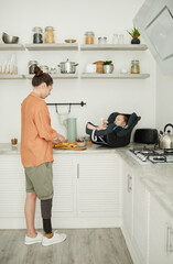 Vertical full length portrait of modern single mother with disability cooking in kitchen with baby in carrier, copy space