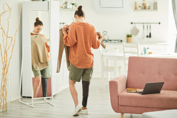 Back view portrait of modern woman with disability looking in mirror while trying on clothes in cozy home interior, copy space