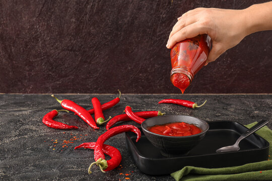 Woman Pouring Chili Sauce From Bottle In Bowl On Dark Background