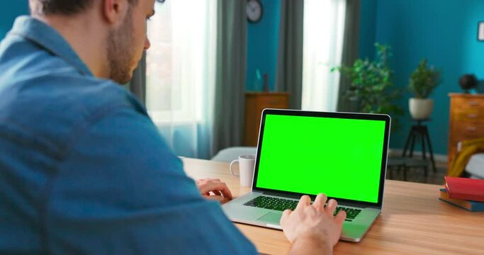 Over the Shoulder Shot of Man uses Laptop with Green Mock-up Screen. In the Background Cozy Living Room.