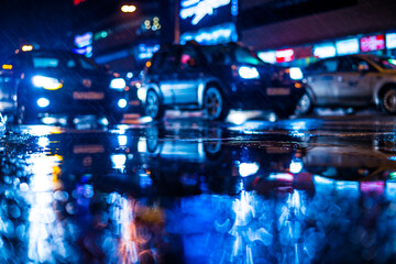 Rainy night in the big city, dense traffic at a busy intersection in the light of shop windows. View from puddles on the pavement level