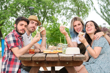 Group of happy friends having fun, drinking and eating sandwiches in a park looking at camera. Picnic on a sunny summer day.