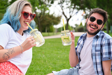 Two friends laughing and drinking refreshing drinks in the park having picnic on a sunny summer day.