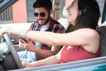 Two friends travel together driving a car, using the smartphone and laughing. Summer adventure.
