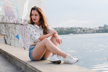 Fototapeta na wymiar Girl in shorts and sneakers walks by a large lake. Girl in short denim shorts on the waterfront of a city lake.