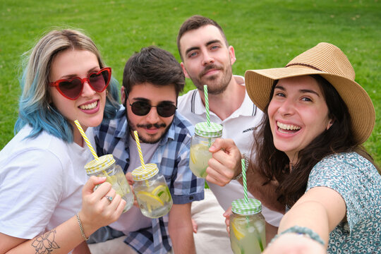 Group of happy friends smiling and taking a selfie while drinking refreshing drinks in the park having picnic on a sunny summer day.