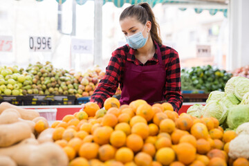 Obraz premium Young saleswoman in a protective mask, working in a store during the pandemic, lays out tangerines on the counter