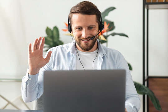 Smiling Indian man in headset with a microphone sit at desk at home office look at laptop screen talk on video call, conversation on computer, online conference or education, greeting