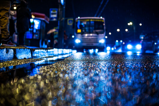Rainy night in the big city, people standing at the bus stop waiting for the bus. View from the level of asphalt