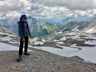 Boy with sunglasses, hat and backpack stands in front of a snow-covered valley in the high mountains on the Zugspitze in Germany with a cloudy sky in the background.