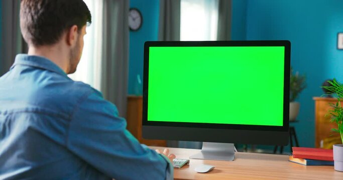 Man Uses all in one computert with blank Screen. Zoom in On a Modern Personal Computer with Mock-up Green Screen Display Standing on the Desk of the Cozy Home Office.