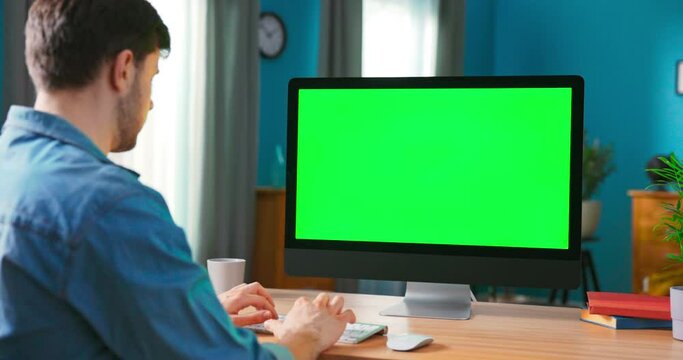 Over the Shoulder Shot of Man Typing on a keyboard computer with blank screen. Zoom in On a Modern Personal Computer with Mock-up Green Screen Display Standing on the Desk of the Cozy Home