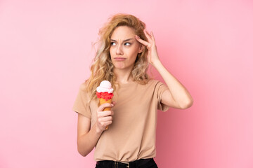 Young blonde woman holding a cornet ice cream isolated on pink background having doubts and with...