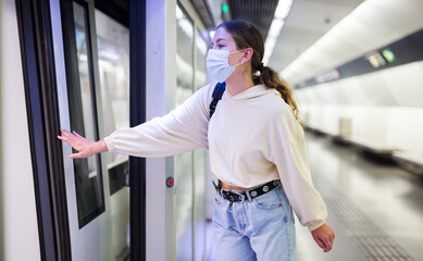 Obraz premium Confident girl in a protective mask, standing on the platform of a subway station during a pandemic, is about to enter ..the train