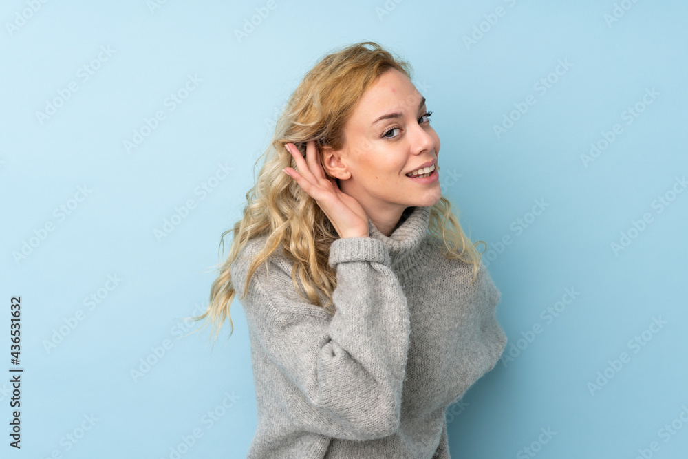 Poster Young blonde woman wearing a sweater isolated on blue background listening to something by putting hand on the ear