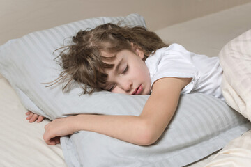 A little girl sleeps sweetly under a gray blanket, on a white sheet and hugs a big gray pillow with her hands.
