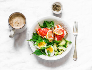 Cappuccino and a bowl of green salad, avocado, boiled egg and sandwiches with rye bread, cream cheese and salmon on a light background, top view. Delicious and healthy breakfast, brunch