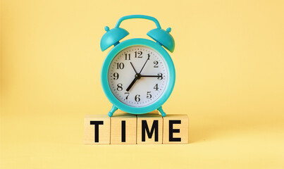 On a light yellow background, wooden cubes with the word TIME and with a blue alarm clock from above