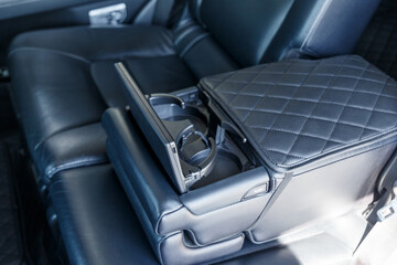 Close-up of the rear armrest with cup holders. The back row of an expensive car with an unfolded armrest. Car interior of luxury leather interior trim with white diamond stitching. Car detail