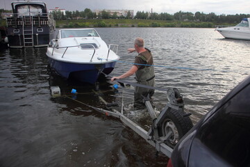 Blue white motor boat launch, a European man puts a boat on the car trailer from water on shipway...