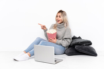 Teenager blonde girl eating popcorn while watching a movie on the laptop pointing finger to the side and presenting a product