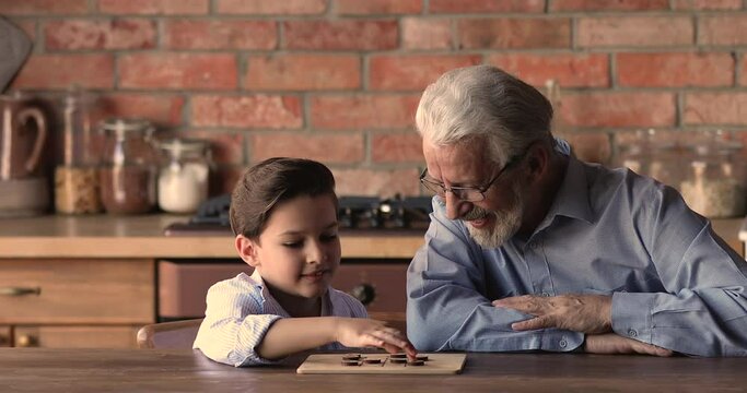 Grey-haired grandpa play checkers with small grandson, family spend free time together sit at table in kitchen have fun enjoy draughts board game. Children development, intellectual home hobby concept