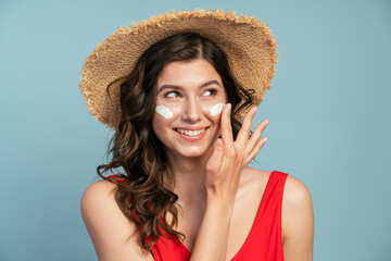 Smiling female wearing swimsuit with straw hat and smearing sunblock lotion on face looking away on blue background. Skin protection concept
