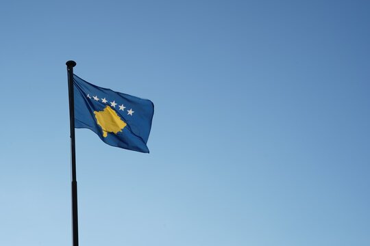 Kosovo flag hoisted on a flagpole waving in the wind. Low angle view against blue sky with plenty of copy space.  - Powered by Adobe