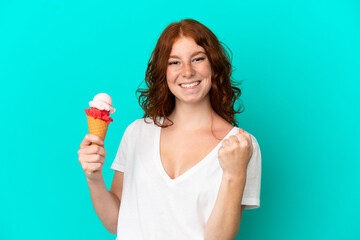 Teenager reddish woman with a cornet ice cream isolated on blue background celebrating a victory in...