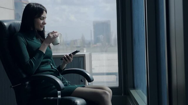 Beautiful woman drinks tea and looks at the phone in the office. Portrait of a business woman with a cup of coffee, in the office of a business center near a large panoramic window