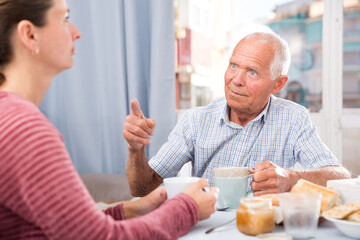 Senior man talking with young sad daughter at table with food and pointing to mistakes