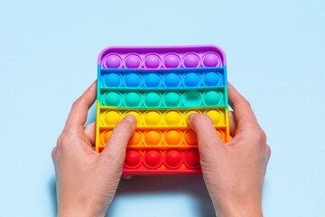 Kid's hands is holding a rainbow Pop It Toy against a blue background. Trendy colorful fidget...