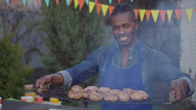 Portrait of happy smiling African American man posing with burger patties on bbq grid outdoors. Joyful handsome young chef with toothy smile bragging roasted grilled meat on picnic barbecue